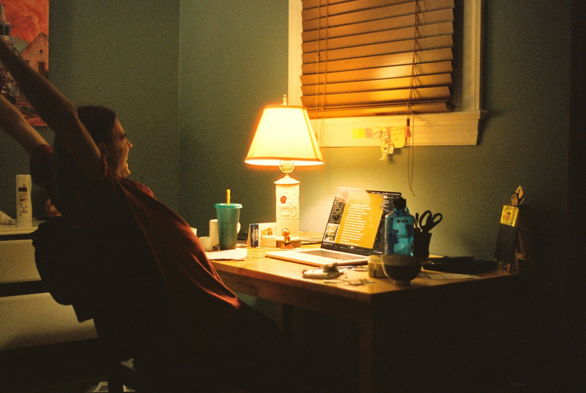 A person stretching at a desk lit by a lamp at the end of a long day